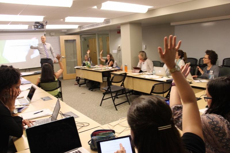 Participant in foreground raises hand while LCIRT presenter speaks to students around tables in Landmark College classroom